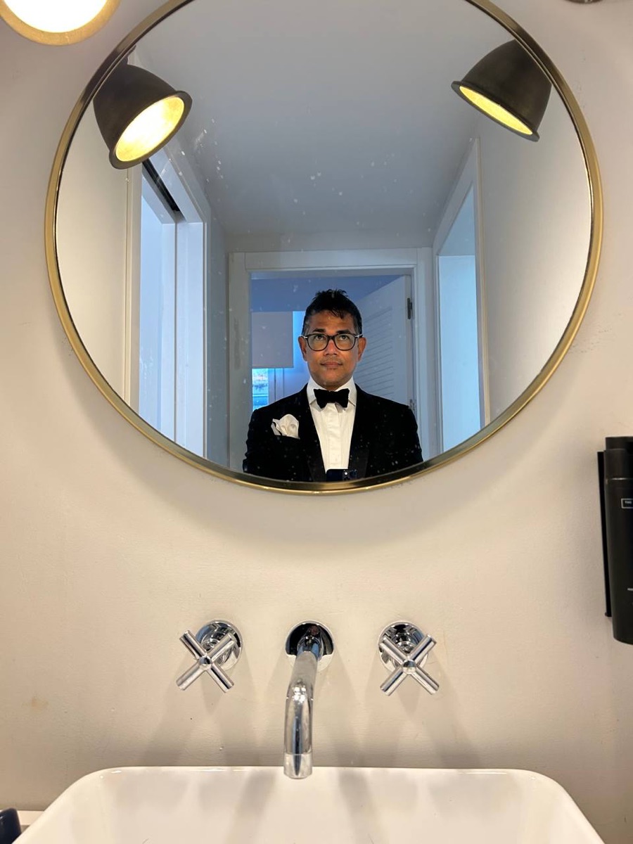 A man in black tie, photographed in a round gold mirror above a bathroom sink